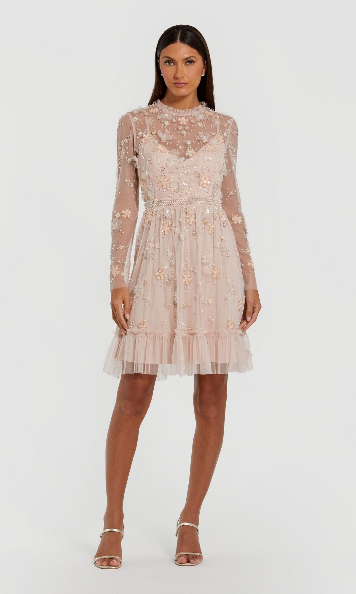 A woman wears the Mac Duggal 9140 short blush wedding guest dress featuring long sleeves, sheer nude fabric, floral embroidery, tulle skirt, and pearl details, paired with strappy heeled sandals.